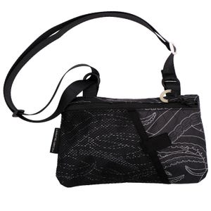 May include: Black crossbody bag with a white octopus tentacle design. The bag has a mesh pocket, a black adjustable strap, and a zipper closure. The bag is designed for carrying essentials.