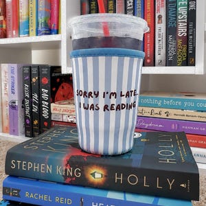 May include: A clear plastic iced coffee cup with a red straw, encased in a light blue and white vertical striped neoprene sleeve. The sleeve features the black text, "SORRY I'M LATE... I WAS READING," printed clearly on the front surface.