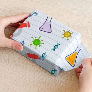 May include: A wrapped gift box with a science-themed pattern. The white paper features illustrations of beakers, DNA strands, and viruses in various colors. The box is being held by hands on a light wood surface.