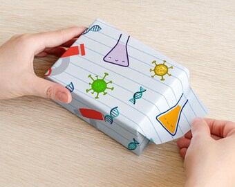 May include: A wrapped gift box with a science-themed pattern. The white paper features illustrations of beakers, DNA strands, and viruses in various colors. The box is being held by hands on a light wood surface.