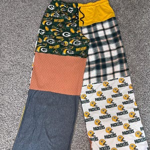 May include: Wide-leg pants featuring a patchwork design with various fabrics. One side displays Green Bay Packers logos on green, a solid rust-colored section with white dots, and a dark gray fabric. The other side has a yellow waistband, plaid, and white fabric with Packers helmet graphics.