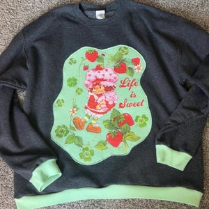 May include: A dark gray crewneck sweatshirt with light green ribbed cuffs and hem. The center features a large, irregular light green patch with a graphic of a character in a strawberry hat holding a pink animal, surrounded by strawberries and the text Life is Sweet.