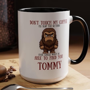 May include: White coffee mug with a black interior and handle. The mug features the text "DON'T TOUCH MY COFFEE I'LL SLAP YOU SO HARD EVEN GOOGLE WON'T BE ABLE TO FIND YOU TOMMY" and a cartoon illustration of a Bigfoot.