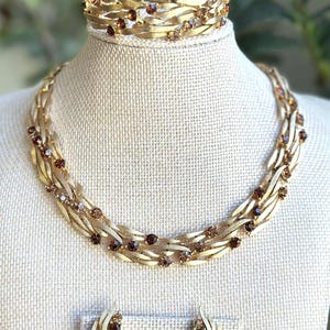 May include: A vintage gold-toned jewelry set featuring a necklace, bracelet, and earrings. The necklace and bracelet have a layered design with brown gemstone accents. The earrings are a matching style. The jewelry is displayed on a mannequin.
