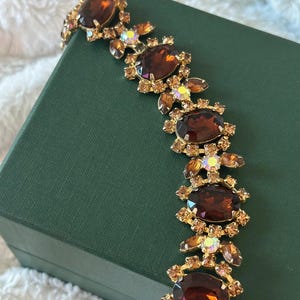 May include: A vintage bracelet featuring large, oval, dark amber-colored rhinestones set in a gold-tone metal. Smaller, iridescent stones and amber-colored accents surround the larger stones. The bracelet is displayed on a green box.