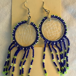 May include: A pair of handmade dreamcatcher earrings. Each earring features a circular frame with a webbed center, surrounded by blue and green beads. Long strands of blue beads with green accents dangle from the bottom.