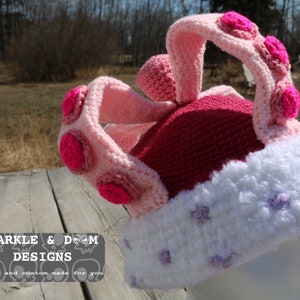 May include: A pink and white crocheted hat with a crown design and purple flower accents. The hat is made with a soft, fluffy yarn and has a unique, whimsical style.