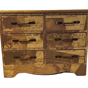 May include: A small, decorative chest of drawers with six drawers. The chest is covered in a collage of sheet music and sepia-toned images. The drawer pulls are dark metal. The top and edges are trimmed with gold. The chest has a curved base.