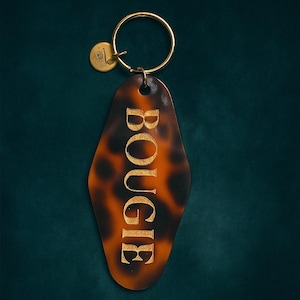 May include: A vintage-style keychain with a tortoiseshell pattern and the word "BOUGIE" in gold lettering. The keychain has a gold ring and a small, round tag. The overall design is reminiscent of retro motel keychains.