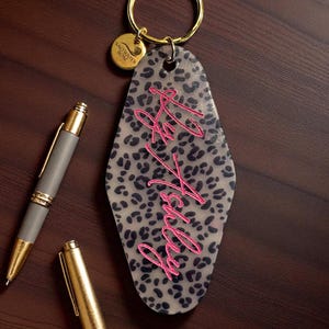 May include: A keychain with a leopard print design and the words "Hey Ashley" in pink script. The keychain has a gold ring and a small gold tag that says "Saltwater Rose". A gray pen and a gold pen are next to the keychain.