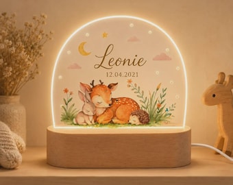 Op de afbeelding: Een boogvormig LED-nachtlampje met houten voet met een aquarelillustratie van een reekalf, een konijn en een egel in het gras. De tekst "Leonie" en "12.04.2021" staat boven de dieren, geaccentueerd door een halve maan en sterren.