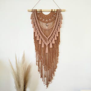 May include: A handcrafted macrame wall hanging in shades of brown and tan. The bohemian-style decor features intricate knotting, tassels, and a crescent moon charm. It is suspended from a natural wooden dowel, adding a touch of rustic charm.
