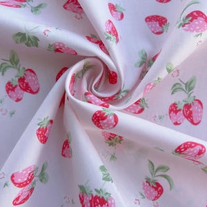 May include: White fabric featuring a repeating pattern of pink and red strawberries and green leaves. The strawberries have a dotted texture and are in various stages of ripeness. The fabric is folded, creating shadows and depth.