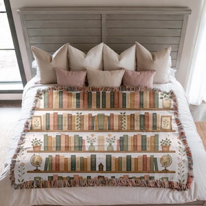 Puede incluir: Una manta decorativa con un diseño de estantería. La manta muestra filas de libros en varios colores, junto con plantas en macetas, cuadros enmarcados y un globo terráqueo. Los bordes tienen un ribete con flecos, y la paleta de colores incluye tonos de marrón, verde y crema.