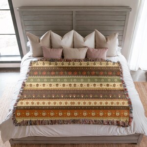 May include: A woven throw blanket featuring horizontal bands of autumn-themed patterns including pumpkins, acorns, mushrooms, leaves, and coffee mugs in shades of orange, brown, green, and cream, finished with a fringed edge on the top and bottom.