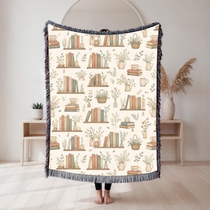 Puede incluir: Manta crema con un patrón repetido de libros, estanterías, flores y tazas de té. La manta tiene un fleco azul oscuro en los bordes. El diseño presenta colores apagados de verde, marrón y naranja.