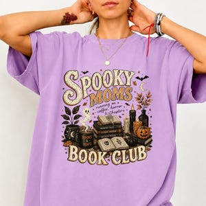 Puede incluir: Una camiseta de color morado claro con un estampado gráfico que incluye el texto "SPOOKY MOMS BOOK CLUB" y "running on coffee, horror & chapters". El diseño muestra ilustraciones de libros apilados, un fantasma, una calabaza, velas y una taza.
