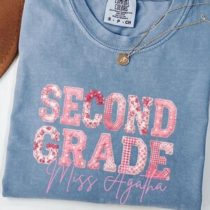 Puede incluir: Una camiseta azul de manga corta con cuello redondo y etiqueta Comfort Colors. El frente presenta el texto SECOND GRADE en letras de bloque rosas con patrones, con el nombre Miss Agatha escrito en cursiva rosa debajo del texto.