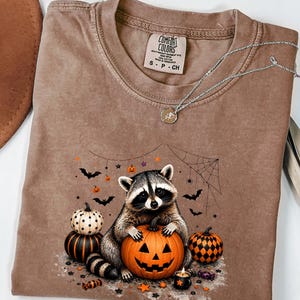 Puede incluir: Una camiseta marrón de cuello redondo con un gráfico de un mapache sosteniendo una calabaza de Halloween, rodeado de calabazas pequeñas, murciélagos, una telaraña y estrellas. La prenda incluye una etiqueta Comfort Colors y un collar de plata.