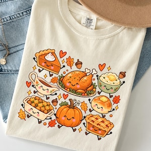 Puede incluir: Una camiseta de color crema con un estampado gráfico de alimentos de Acción de Gracias sonrientes, que incluye un pavo asado, pastel de calabaza, puré de patatas, relleno y una calabaza, rodeados de pequeños corazones y hojas de otoño.