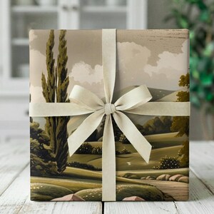Puede incluir: Una caja de regalo cuadrada envuelta en papel con una ilustración de paisaje de colinas verdes, cipreses altos y un cielo nublado. El paquete está atado con una cinta de satén de color crema dispuesta en un lazo centrado en la parte frontal.