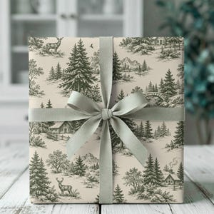 May include: A square gift box wrapped in cream-colored paper featuring a dark green toile-style print of a winter forest landscape with deer, pine trees, mountains, and cabins. The box is tied with a solid light green fabric ribbon bow.
