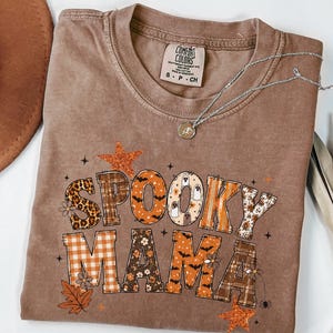 Puede incluir: Una camiseta marrón de manga corta con cuello redondo y etiqueta de la marca Comfort Colors. El frente presenta el texto SPOOKY MAMA en letras de bloque con varios patrones que incluyen estampado de leopardo, cuadros naranjas, fantasmas y murciélagos.
