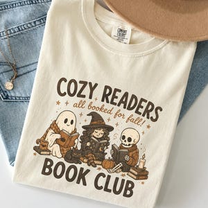 Puede incluir: Una camiseta de color crema con un estampado gráfico marrón con el texto COZY READERS all booked for fall! BOOK CLUB. La ilustración muestra a un fantasma, una bruja y un esqueleto leyendo libros, acompañados por un gato negro, una calabaza y una vela.