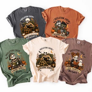 Op de afbeelding: Vijf t-shirts in antraciet, oranje, saliegroen, crème en mauve met grafische prints van skeletten die boeken lezen. Elk ontwerp bevat de tekst SKELETON CREW, READING BONES, DADDY BONES of BIG BONES, samen met namen zoals Lucas, Masson, Dave en Harper.