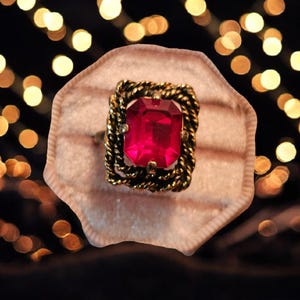 May include: A vintage-style ring with a large, rectangular, ruby-red gemstone. The ring has a gold-colored, twisted rope design around the gemstone. The ring is presented in a light pink ring box.