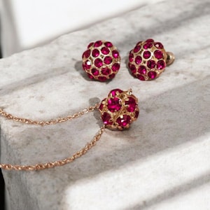 May include: A gold-toned jewelry set featuring a necklace and matching earrings. Each piece has a spherical design encrusted with vibrant red gemstones. The set is displayed on a textured, light-colored surface.
