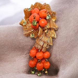 Puede incluir: Broche vintage dorado con cuentas naranjas y detalles de pedrería verde. El broche tiene un diseño texturizado único con forma de abanico y elementos colgantes de cuentas. Un accesorio elegante para una chaqueta o blusa.