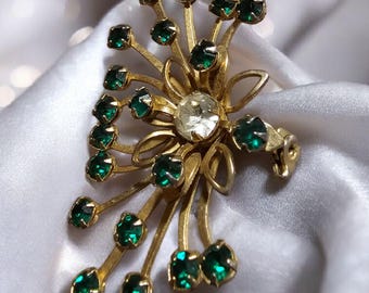 May include: Vintage gold-tone brooch with a floral design. The brooch features a central clear rhinestone surrounded by gold petals and stems adorned with emerald green rhinestones. A classic piece of jewelry.