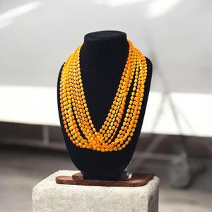 May include: A multi-strand necklace featuring numerous small, oval-shaped beads in a vibrant, translucent orange hue. The beads are strung together in several layers, creating a voluminous, cascading effect that drapes across the neck area.