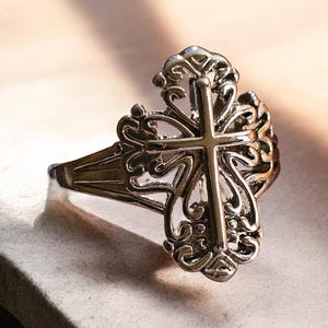 May include: Silver-toned ring featuring a detailed cross design. The ring has an ornate, filigree-style setting around the cross, with a polished finish. The cross is the central focus, with a classic Christian symbol.