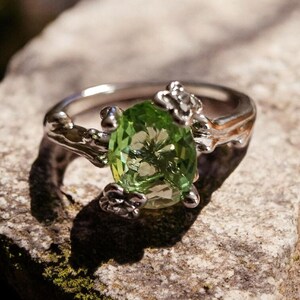 May include: A silver ring featuring a large, oval-cut, light green gemstone. The ring's band has a textured, branch-like design, with small decorative elements around the gemstone. The ring is set on a rough, grey stone surface.