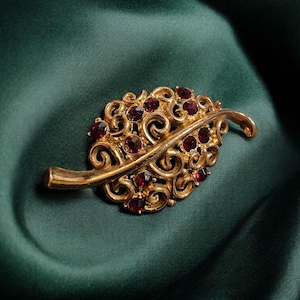 Op de afbeelding: Goudkleurige broche in de vorm van een blad met kleine, ronde, rode edelstenen. De broche heeft ingewikkeld krulwerk en een gebogen steel. De achtergrond is een donkergroene, zijdeachtige stof.