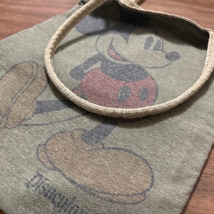 May include: A green tote bag featuring a large graphic of Mickey Mouse. The illustration shows Mickey in his classic pose, with red shorts and gray shoes. The bag has a beige handle and the word "Disneyland" is printed at the bottom.