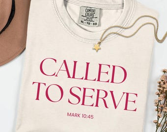 Può includere: T-shirt color crema con le parole "CALLED TO SERVE" in grassetto, colore magenta. Il testo "MARK 10:45" è in basso. Sono visibili anche una collana con pendente a stella dorata e un cappello marrone.