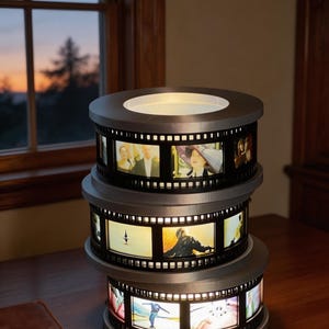 May include: A three-tiered lamp with a film reel design. Each tier displays illuminated film stills. The lamp has a silver-grey metal frame and sits on a wooden surface. The background shows a window with an outdoor view.