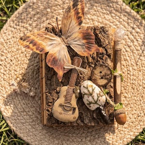 May include: A rectangular bark-covered journal featuring a large brown butterfly, a small fabric guitar, a dark wooden button, a white embroidered mushroom, and a twig pen with a crystal topper. The text 100% NATURAL & HANDMADE is visible.
