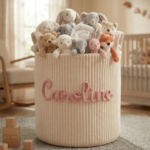 Puede incluir: Un cesto de almacenamiento de pana beige lleno de animales de peluche. El cesto tiene el nombre bordado en rosa, "Caroline", y está lleno de conejos, elefantes, osos y un zorro de peluche. El cesto tiene dos asas y está sobre una alfombra gris.