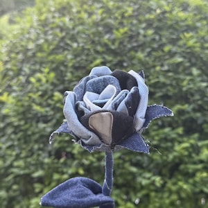 Pode incluir: Uma rosa de ganga artesanal. As p&eacute;talas s&atilde;o feitas de v&aacute;rias tonalidades de ganga azul, com um centro castanho e preto. O caule e a folha tamb&eacute;m s&atilde;o de ganga, criando um design floral &uacute;nico.