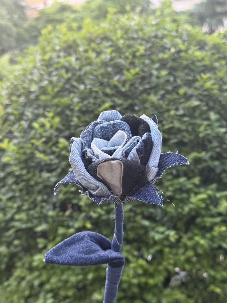Pode incluir: Uma rosa de ganga artesanal. As p&eacute;talas s&atilde;o feitas de v&aacute;rias tonalidades de ganga azul, com um centro castanho e preto. O caule e a folha tamb&eacute;m s&atilde;o de ganga, criando um design floral &uacute;nico.