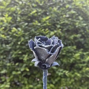 Pode incluir: Uma rosa de ganga artesanal com caule e p&eacute;talas azuis. As p&eacute;talas da flor s&atilde;o feitas de tecido de ganga azul escuro com bordas desfiadas, dando-lhe uma apar&ecirc;ncia r&uacute;stica. O caule &eacute; de uma cor azul correspondente. O fundo &eacute; uma folhagem verde desfocada.