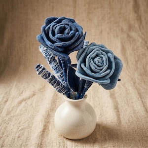 Pode incluir: Um pequeno vaso de cer&acirc;mica branca cont&eacute;m um buqu&ecirc; de rosas de ganga. As rosas s&atilde;o feitas de v&aacute;rias tonalidades de ganga azul, com formas detalhadas de p&eacute;talas. Os caules e as folhas tamb&eacute;m s&atilde;o de ganga. O vaso est&aacute; sobre um tecido de tom neutro.