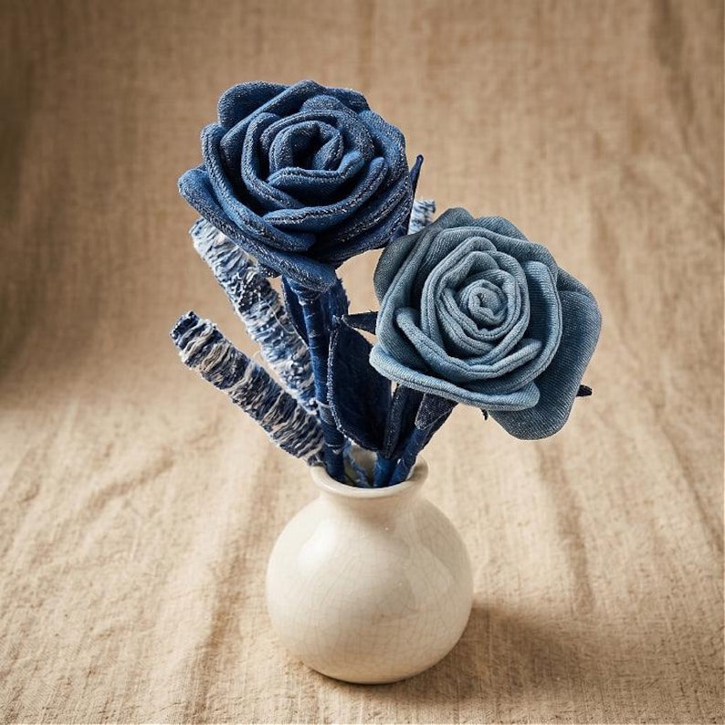Pode incluir: Um pequeno vaso de cer&acirc;mica branca cont&eacute;m um buqu&ecirc; de rosas de ganga. As rosas s&atilde;o feitas de v&aacute;rias tonalidades de ganga azul, com formas detalhadas de p&eacute;talas. Os caules e as folhas tamb&eacute;m s&atilde;o de ganga. O vaso est&aacute; sobre um tecido de tom neutro.
