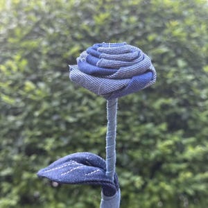 Pode incluir: Uma rosa de ganga feita &agrave; m&atilde;o. A cabe&ccedil;a da flor tem um padr&atilde;o azul e branco, com um caule e uma folha. A rosa &eacute; feita de tecido de ganga reciclado. O fundo &eacute; um arbusto verde desfocado.