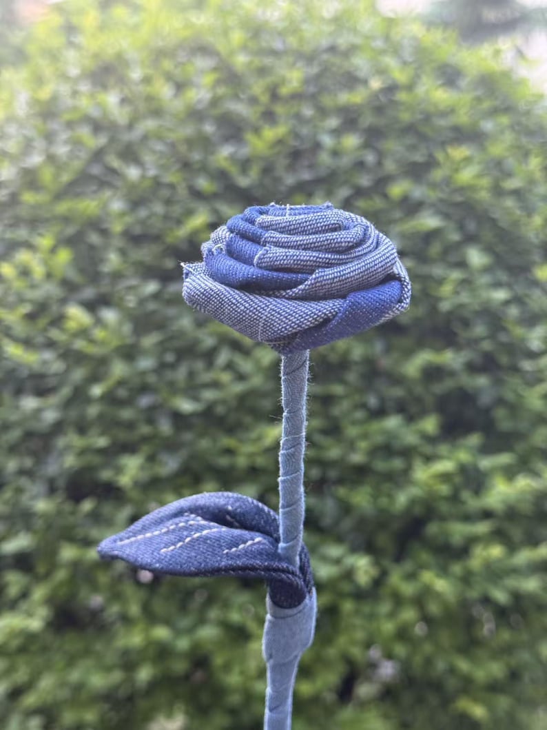 Pode incluir: Uma rosa de ganga feita &agrave; m&atilde;o. A cabe&ccedil;a da flor tem um padr&atilde;o azul e branco, com um caule e uma folha. A rosa &eacute; feita de tecido de ganga reciclado. O fundo &eacute; um arbusto verde desfocado.