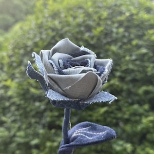 Pode incluir: Uma rosa de ganga artesanal com caule e folha. As p&eacute;talas da flor s&atilde;o feitas de v&aacute;rias tonalidades de ganga azul, criando uma apar&ecirc;ncia texturizada e &uacute;nica. O caule e a folha tamb&eacute;m s&atilde;o feitos de ganga.